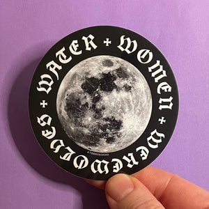 Kristin's hand holds a 3-inch black and white circular sticker depicting the moon against a purple background. The words "water, women, werewolves" in a gothic typeface form a ring around the perimeter.