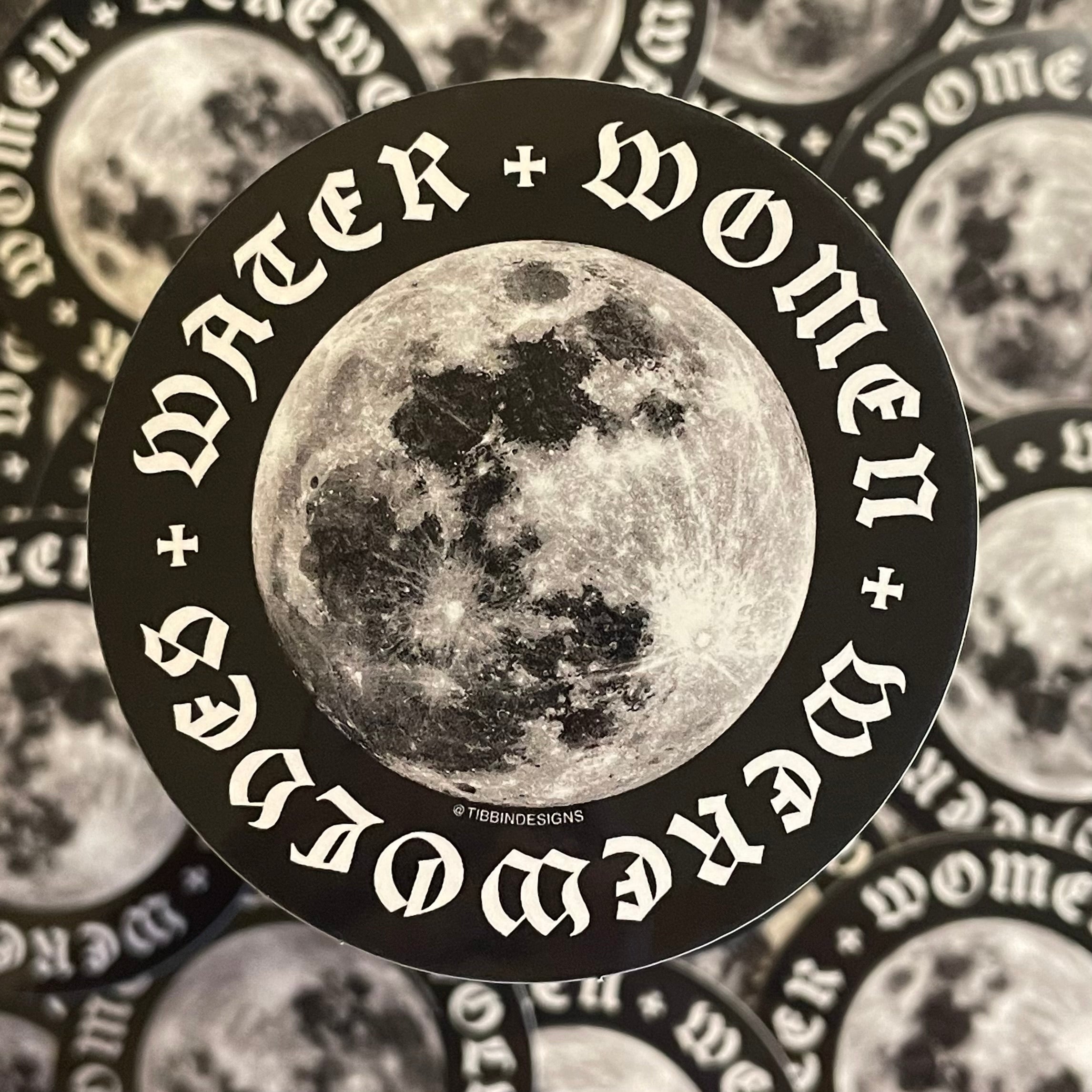 A 3-inch black and white circular sticker depicting the moon sits against a focus-blurred collage of the same sticker. The words "water, women, werewolves" in a gothic typeface form a ring around the perimeter.