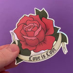 Load and play video in Gallery viewer, A video shows the holgraphic effect of a sticker of a red rose drawn in the traditional American tattoo style with a tattoo banner that says "Love Is Love" in a gothic font. The red rose has pink petals as accents.
